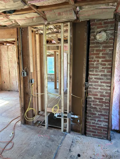 Interior under construction with exposed stud framing, brick chimney, and rough-in electrical and plumbing work.
