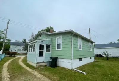 Side view of a small green house with a gambrel roof, light blue porch addition, and white foundation.
