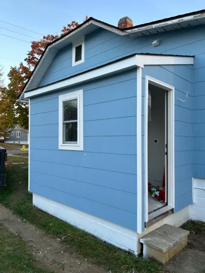 Side view of a blue-sided home addition under construction with an open door showing unfinished interior work.