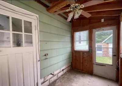 Unfinished porch room with exposed beams, green lap siding, and a door leading to a wooded yard.