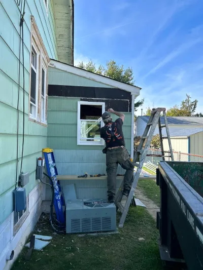 A contractor on a ladder repairs siding on a house exterior near an HVAC unit and dumpster.