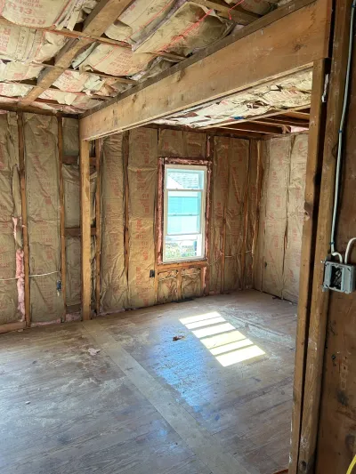 A gutted room mid-renovation with exposed stud framing, insulation, and bare subfloor, lit by a single window.
