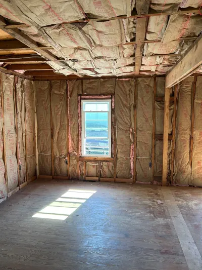 Room mid-renovation with exposed wall studs, ceiling joists, and fiberglass insulation, lit by a single window.