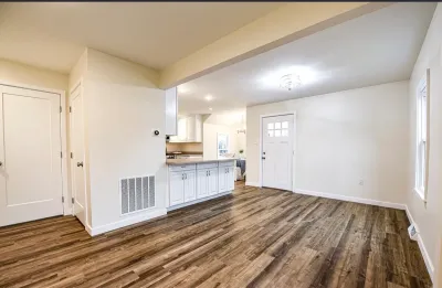Open living area with wide-plank flooring flows into a white shaker-cabinet kitchen and entry door.