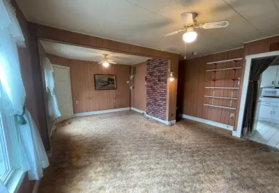 Dated living room with wood paneling, brick accent wall, worn carpet, and ceiling fans before renovation.