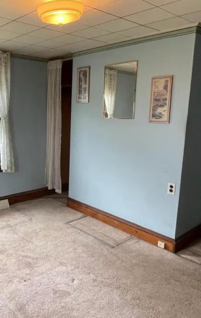 Empty bedroom before renovation with blue walls, worn carpet, and a curtained closet opening.