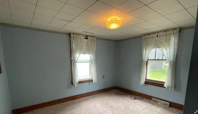 Empty bedroom with light blue walls, dated drop ceiling, carpet flooring, and two curtained windows.