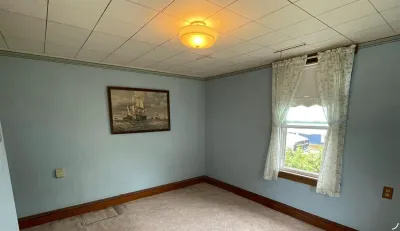 Empty blue bedroom with carpet, dated drop ceiling, and a window with floral curtains.