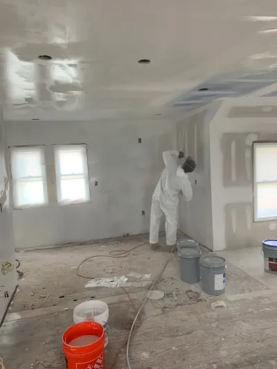 A worker in a protective suit sands drywall seams in a room mid-renovation with exposed subfloor and rough electrical wiring.