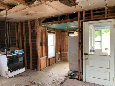A gutted kitchen mid-renovation with exposed wall studs, ceiling joists, and appliances left in place.