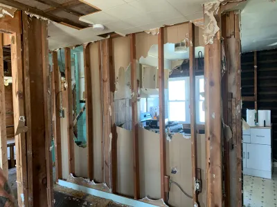 Kitchen mid-demolition with exposed wood stud walls, wiring, and a partially intact cabinet near a window.