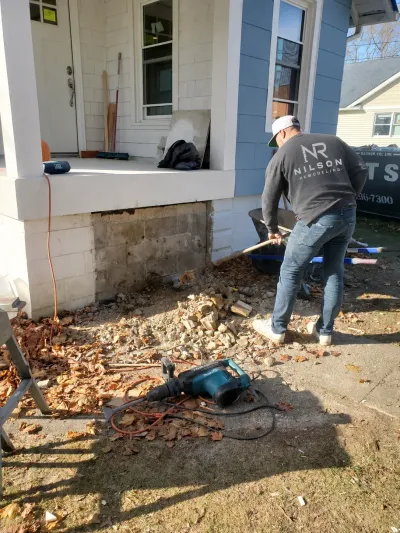 Worker clears rubble from a demolished foundation wall beneath a front porch during remodeling.