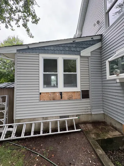 Side of a house mid-repair with vinyl siding removed below two windows, exposing plywood sheathing.