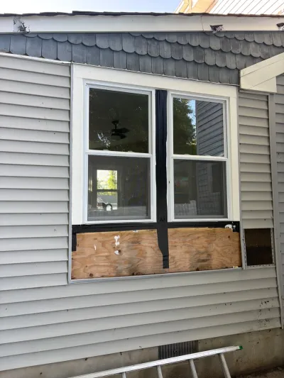 Exterior wall during repair with siding removed below two windows, exposing plywood sheathing and a ladder against the founda
