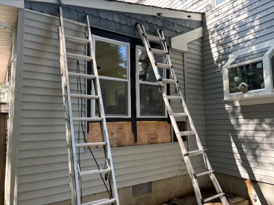 Two ladders lean against a house exterior near a window with a plywood repair patch underneath it.