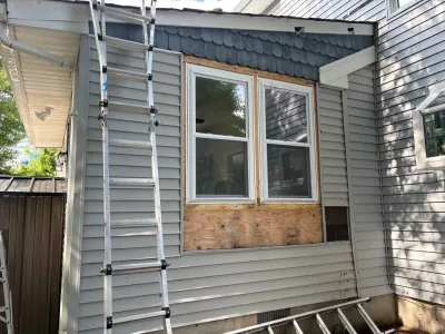 Exterior wall of a house with vinyl siding removed around a window, exposing plywood, and a ladder set up for repair work.