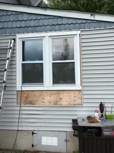 Exterior wall with vinyl siding removed below a window, exposing plywood, with a ladder leaning nearby.