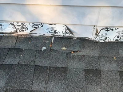 Close-up of damaged roof shingles and torn house wrap exposed beneath vinyl siding at a roofline.