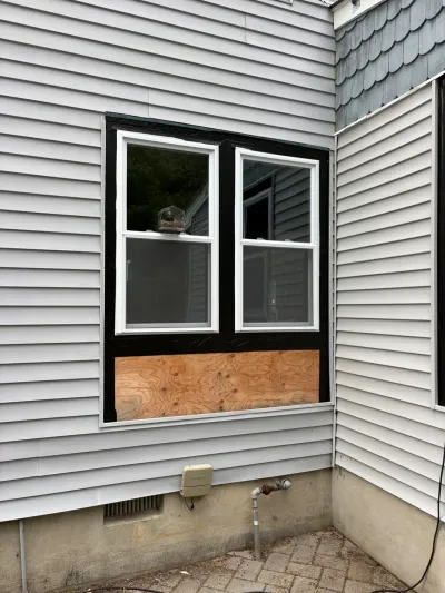 Exterior wall mid-renovation with a new vinyl window, black flashing tape, and plywood infill below the sash.