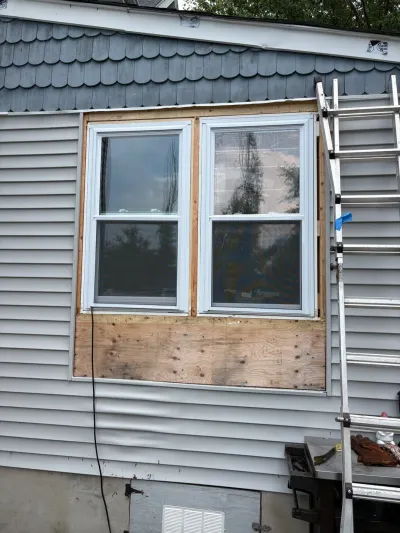Two new vinyl windows installed with surrounding wood framing and plywood patch, ladder set up alongside for siding work.