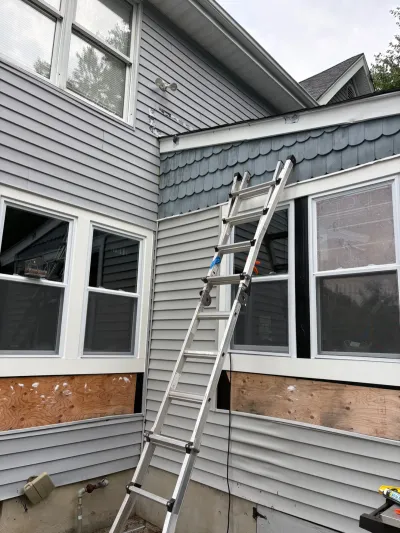 Ladder leans against a house under repair, with plywood patches where siding has been removed.