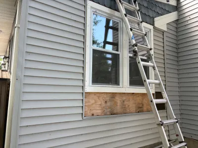 Exterior wall with vinyl siding removed below a window, exposing plywood, with a ladder set up for repair work.