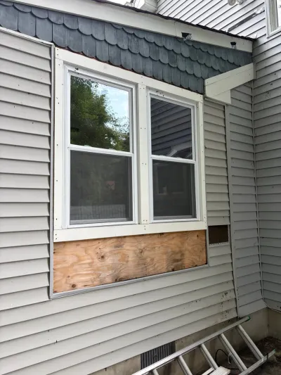 House exterior mid-repair with vinyl siding removed below a window, revealing plywood sheathing.