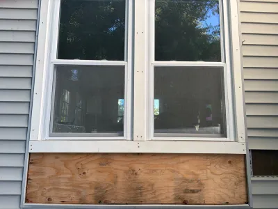 Exterior wall with two windows and a section of siding removed, exposing plywood sheathing below.