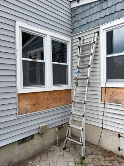 Exterior wall with vinyl siding removed below two windows, exposing plywood, with a ladder set up for repair work.