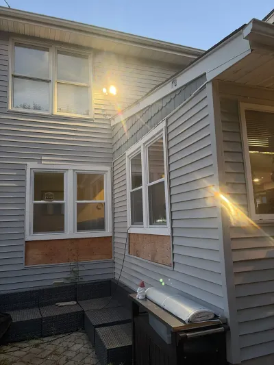 Exterior of a house mid-repair with plywood patches below windows and vinyl siding, lit by sunset.