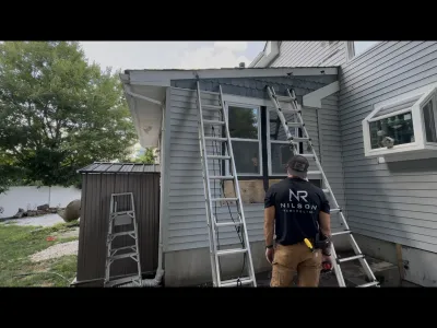 Contractor stands beside ladders at a house exterior where vinyl siding has been removed for repair.