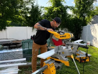 A worker cuts a white trim board on a miter saw in a backyard enclosed by a white vinyl fence.