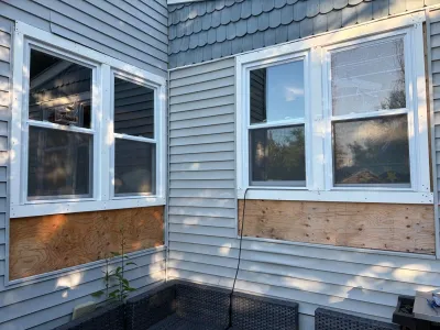 Exterior corner of a house with vinyl siding removed below the windows, exposing plywood sheathing during repairs.