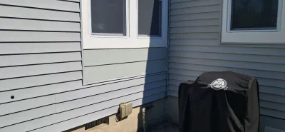 Exterior corner of a house with gray vinyl siding showing a mismatched patched section near two windows.