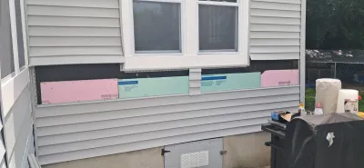 Section of house siding removed to reveal pink and teal foam insulation panels below a window.