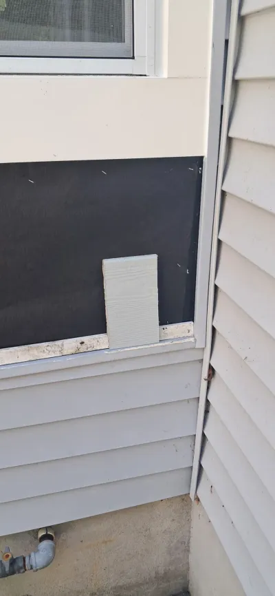 Exterior wall mid-renovation showing exposed black house wrap above vinyl siding and a siding sample piece.