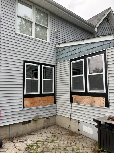 Rear corner of a house mid-renovation with new windows installed and plywood still covering the lower window openings.