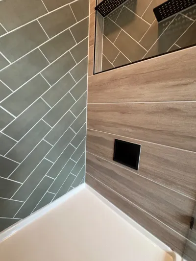 Close-up of a shower corner showing sage herringbone tile meeting wood-look plank tile with a black rainfall shower head.
