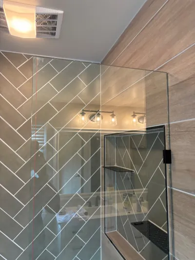 Herringbone-tiled shower with frameless glass door and matte black tiled storage niche beside a wood-look accent wall.
