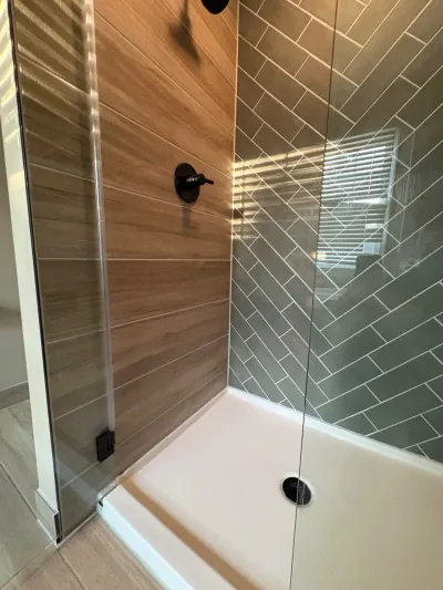 A walk-in shower with wood-look tile on one wall and herringbone grey tile on another, framed by frameless glass.