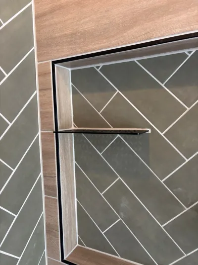 Close-up of a shower corner with sage herringbone tile, wood-look tile trim, and a black-framed glass door.