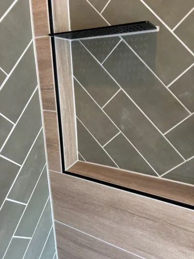 Close-up of a shower corner with green herringbone tile, wood-look accent tile, and a black-framed glass shelf.
