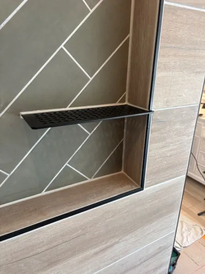 Close-up of a tiled shower niche with a black metal shelf and herringbone sage green tile interior.