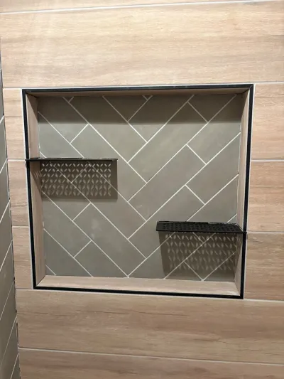 A tiled shower niche with a herringbone pattern and two black metal shelves, set into wood-look wall tile.