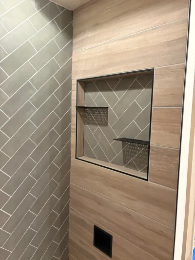 Close-up of a tiled shower wall with a wood-look herringbone pattern and a black-framed tile niche.