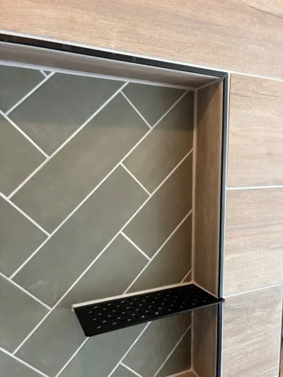 Close-up of a tiled shower niche with herringbone green tile and a black metal shelf insert.