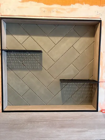 A shower niche mid-installation with herringbone sage tile and two black metal shelves, surrounded by unfinished wall.