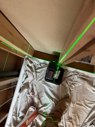 A green laser level projects guide lines across exposed framing in a basement under construction.