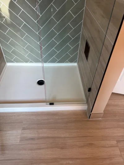 A walk-in shower with sage herringbone tile, a wood-look tile wall, and a frameless glass door.