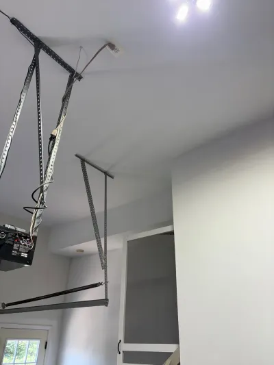 Garage ceiling showing the door opener motor, metal brackets, and a torsion spring, with a stairwell doorway beyond.